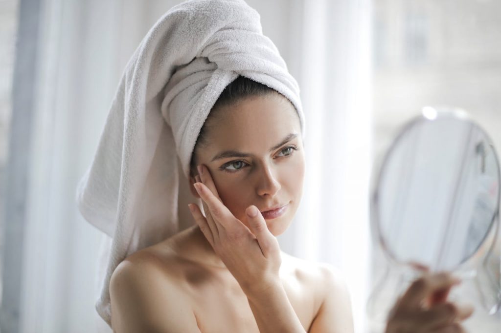 pexels-photo-3754678 Young female with bare shoulders and towel on head holding small mirror and checking skin with it in bathroom