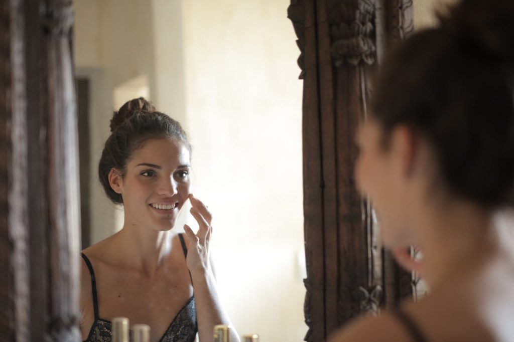 pexels-photo-3785802 Young woman applying skincare and smiling at her reflection in a rustic mirror.